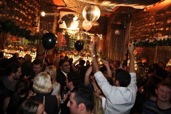 Warm-lit Mumbai bar with disco balls, bottle shelves, and the crowd raising their hands